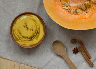 top view pumpkin pureed soup with cream sauce, healthy concept, in clay plate, spices (cinnamon steaks, a star anise, and whole nutmeg), wooden spoon lay on linen cloth, pumpkin with seeds on wooden