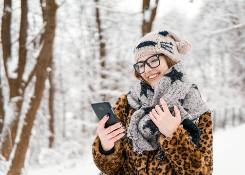 Attractive girl wears glasses holding smartphone in winter snowy park