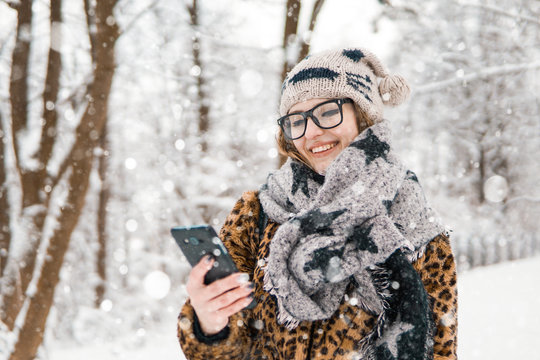 Attractive girl wears glasses holding smartphone in winter snowy park