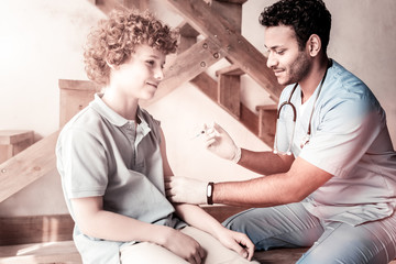 Fototapeta premium Do not worry. Cheerful doctor smiling while getting ready to vaccinate a curly haired boy indoors.