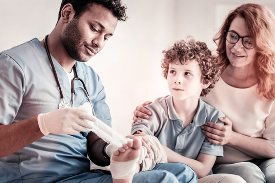 Be careful. Adorable curly haired boy looking at a focused doctor putting a bandage on his foot.