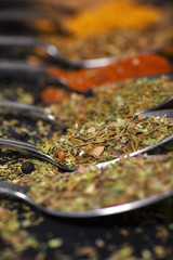 Several steel spoons with spice and herbs lie in the raw on dark background.