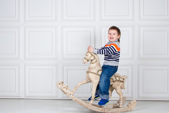 little boy swinging on wooden horse. funny three-year-old boy in jeans and sweater on white background. Carefree childhood
