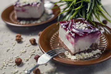 A piece of vanilla cake with chocolate base and cherry glaze decorated with some pieces of white chocolate served in a brown clay dish with silver fork placed on a beige tablecloth. Soft, blur focus.