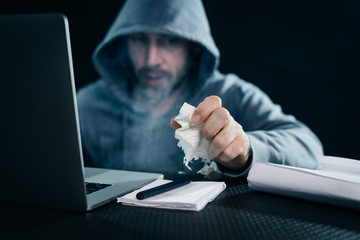a man hacker in a hooded sweatshirt sits in a dark room at a table and crumples a piece of paper