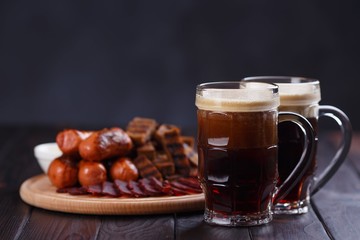 Dark beer and appetizing beer snacks set. Two mugs of stout, wooden board with grilled sausages, basturma and crunchy rye toasts, served with tomato and cheese sauces. Oktoberfest, pub concept