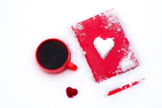 Red cup of coffee or tea with a red book or notebook or photo albums with heart. Snow is coming. Winter concept of Valentine's Day