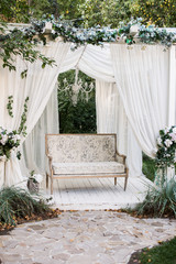In the garden there is a podium on which a beautiful white sofa in the style of Provence or rustic. Above the sofa is an arch with blossoms and flowing white fabric. Decoration for a wedding, scenery