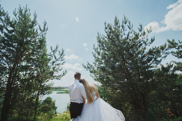 Wedding couple stands near the lake and embrace each other. Blonde bride in puffy dress leans her...