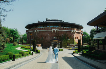 Naklejka premium Wedding couple is walking holding hands near the old ancient citadel castle on sunny day. Bride in puffy dress and groom in light blue suit. Picturesque architecture and view. Green lawn.