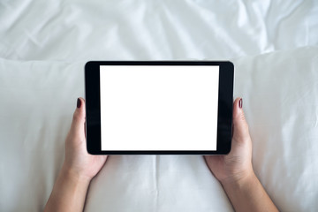 Top view mockup image of a woman holding black tablet pc with blank desktop white screen while sitting on a white bed background