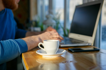 Close-up of men's hands, typing on a laptop, drinking coffee, next to the phone. A cup of coffee is empty. Close-up. Sunlight