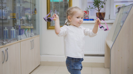 Adorable child blonde girl in ophthalmology clinic plays in hall near glasses store