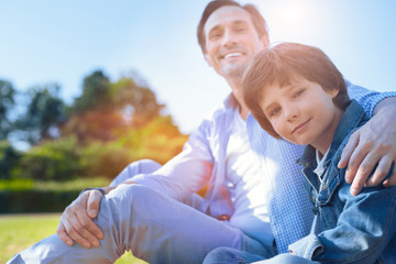 Harmonious relations. Selective focus on a charming youngster looking into the camera with a slight smile on his face while sitting next to his thoughtful father during a male conversation outdoors.