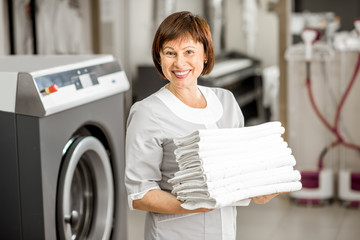 Portrait of a senior washwoman in uniform standing with towels in the hotel laundry - obrazy, fototapety, plakaty