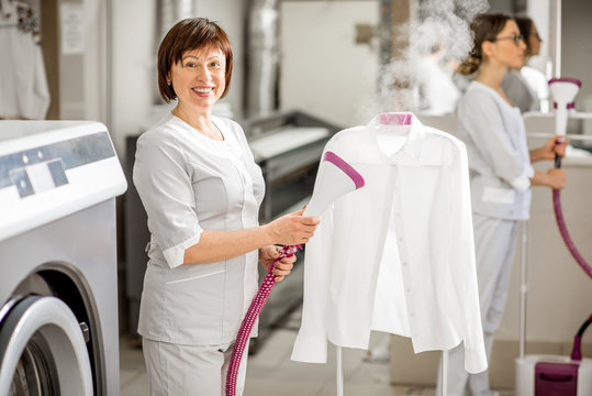 Portrait of a senior washwoman in uniform ironing up a white shirt with vapor machine in the professional laundry