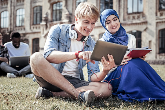 Modern generation. Happy positive cheerful man holding a tablet and using it while sitting on the grass with his friend