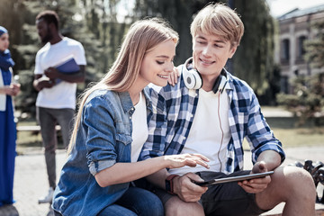 Fototapeta na wymiar Young generation. Happy delighted young woman sitting together with her friend and holing his shoulder while looking at the tablet screen