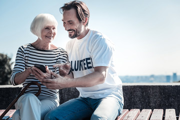 Modern device. Happy nice joyful man pointing at the smartphone and smiling while helping an aged woman to use it