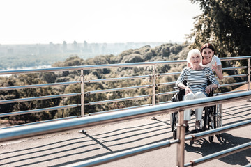 Just look. Positive nice joyful woman standing behind the wheelchair and smiling while pointing with her finger