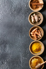 Ingredients for turmeric latte. Ground turmeric, curcuma root, cinnamon, ginger, honeycombs in wooden bowls in row over black texture background. Top view, copy space - obrazy, fototapety, plakaty