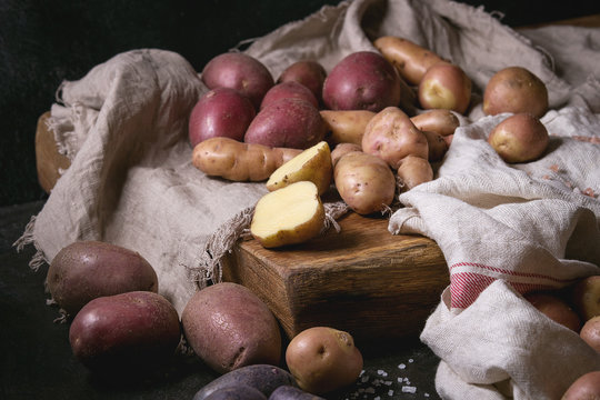 Variety of raw uncooked organic potatoes different kind and colors red, yellow, purple on wooden cutting board with kitchen towels over black table. Dark rustic style