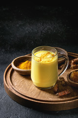 Cup of ayurvedic drink golden milk turmeric latte with curcuma powder on round wooden tray and ingredients above over black texture background. Copy space