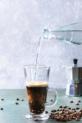 Fizzy iced Coffee espresso with pouring sparkling water from bottle, coffee maker and roasted beans over grey green texture table. Modern drink.