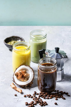 Variety of iced colorful latte drinks. Iced coffee, turmeric and matcha latte cocktails in glass jars with ingredients above over grey green texture background. Copy space