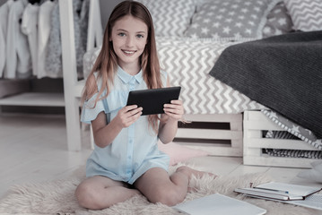 Spare time. Beautiful smiling long-haired girl sitting on the floor in her room and holding a tablet and wearing a blue shirt