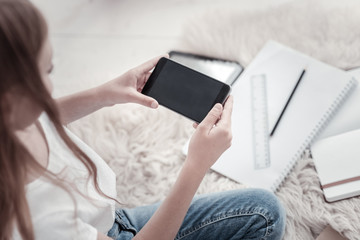 Using phone. Long-haired girl wearing jeans and a white shirt and holding her black phone while sitting on the floor and her notebooks lying near her