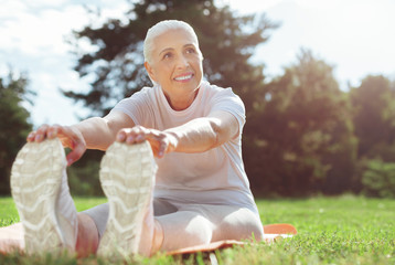 Fototapeta premium Beautiful body. Happy nice well built woman sitting on a yoga mat and stretching while exercising