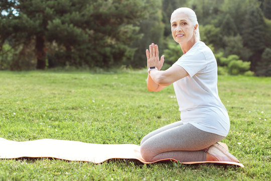 Yoga Practice. Positive Pleasant Aged Woman Sitting On A Yoga Amt And Looking At You While Holding Her Hands Together