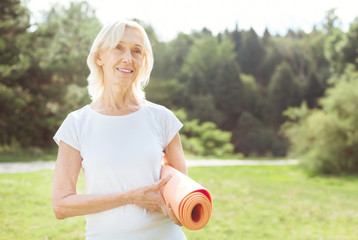 Caring about health. Cheerful positive elderly woman smiling and holding yoga mat while enjoying a...