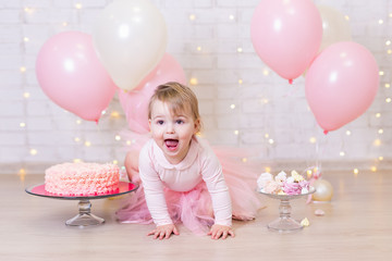 first birthday party and happiness concept - happy little girl with cake and sweets over brick wall background with lights and balloons