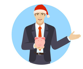 Businessman in Santa hat holding a piggy bank and showing something beside of him