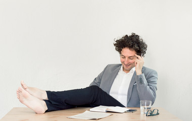 Young, happy, successful Caucasian businessman sitting in office with barefoot on desk and relaxing.