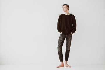 A young man, a student stands against a white wall in the room, dressed in short jeans and a black sweater, barefoot. The guy is a model.