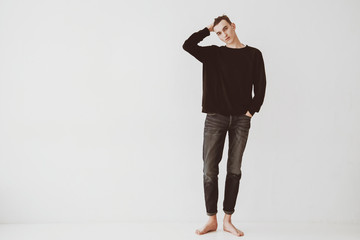 A young man, a student stands against a white wall in the room, dressed in short jeans and a black sweater, barefoot. The guy is a model.