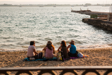 Four girls sitting together on the beach at sunset. Syracuse Ortigia beach, Italy.