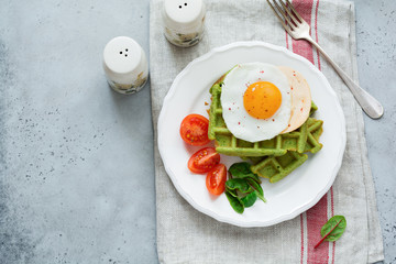 Fried egg with cheese, tomatoes, chard, and waffle with spinach on white ceramic plate on light gray concrete background. Sandwich for breakfast. Selective focus. Top view. Copy space.