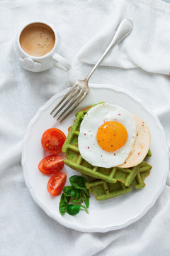 Fried egg with cheese, tomatoes, chard, and waffle with spinach on white ceramic plate on a white rumpled tablecloth. Sandwich for breakfast. Selective focus. Top view. Copy space.