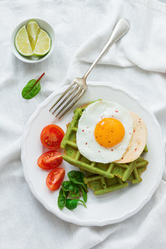 Fried egg with cheese, tomatoes, chard, and waffle with spinach on white ceramic plate on a white rumpled tablecloth. Sandwich for breakfast. Selective focus. Top view. Copy space.