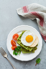 Fried egg with cheese, tomatoes, chard, and waffle with spinach on white ceramic plate on light gray concrete background. Sandwich for breakfast. Selective focus. Top view. Copy space.