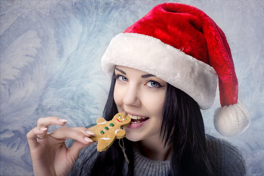 Brunette girl in Santa hat bites the gingerbread man on blue background winter patterns