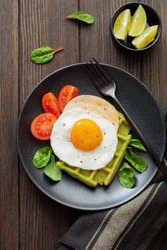 Fried egg with cheese, tomatoes, chard, and waffle with spinach on black ceramic plate on brown old wooden background. Sandwich for breakfast. Selective focus. Top view. Copy space.
