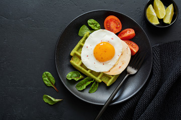 Fried egg with cheese, tomatoes, chard, and waffle with spinach on black ceramic plate on dark concrete background. Sandwich for breakfast. Selective focus. Top view. Copy space.