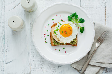 Toast with guacamole sauce from avocado, cheese, fried egg and fresh on white ceramic plate on light   wooden background. Selective focus. Top view.