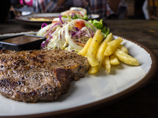 close up of T-bone steak with salt and pepper, golden french fries and green vegetables. on white plate, Fork and knife, wooden table in the restaurant