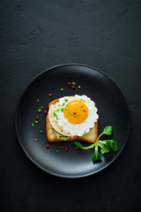 Toast with guacamole sauce from avocado, cheese, fried egg and fresh on black ceramic plate on dark concrete background. Selective focus. Top view.